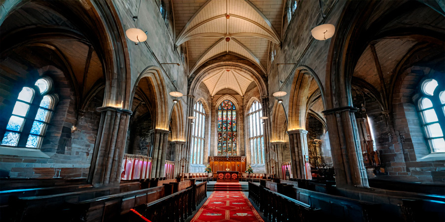 Interior of a historic church with vaulted ceilings, stained glass windows, and an ornate altar at the front. LED lights illuminate the columns, casting a warm glow on the stone walls. A red carpet runs along the center aisle between dark wooden pews.