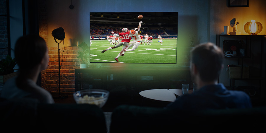 A couple sitting on a couch in a cozy living room, watching a football game on a large television, with a bowl of popcorn on the table in front of them.