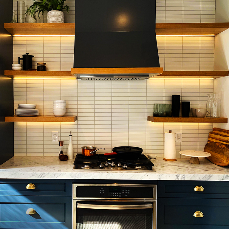 Modern kitchen with blue cabinets, white tiles, and floating shelves lightened with LED strips