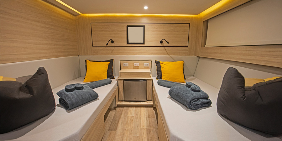  A modern boat cabin featuring LED strip lighting along the ceiling, warm wood finishes, twin beds with yellow pillows, and neatly folded towels for a cozy interior.