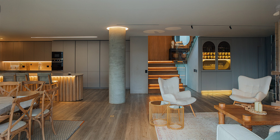 Warm ligModern living and dining area with warm wooden floors and minimalist decor. LED strip lights accentuate a staircase, a ceiling column, and cabinets, creating a cozy ambiance. The space includes light-colored armchairs, a wooden dining table, and built-in shelving with soft lighting.