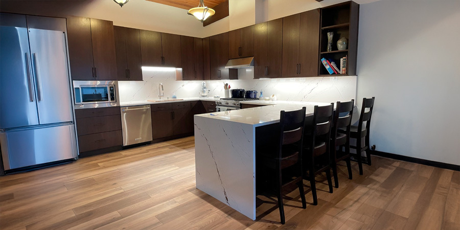 A modern kitchen with dark wood cabinets, a large white marble countertop island, and under-cabinet lighting that illuminates the workspace.