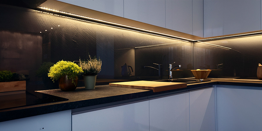 Close-up of a modern kitchen countertop with LED strip lighting under the cabinets, illuminating a dark backsplash. The countertop features a wooden cutting board, a potted plant, and a sink with a chrome faucet.