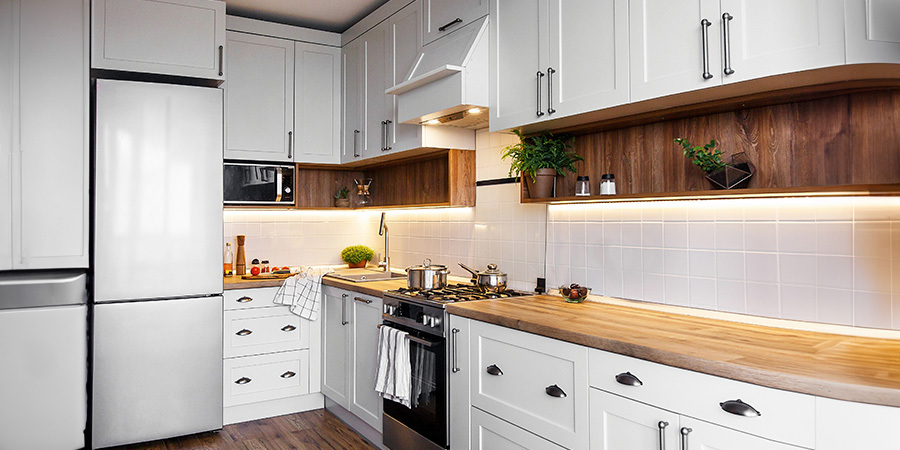 A modern kitchen featuring white cabinets, warm lighting highlighting wood accents in shelving and beneath the upper cabinets, stainless steel appliances, and a light wood floor.