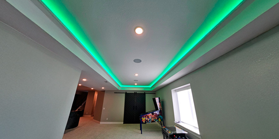 A stylish game room featuring recessed ceiling lighting with green LED strips, a pinball machine, arcade cabinet, and a cozy seating area, enhancing the ambiance with vibrant illumination.