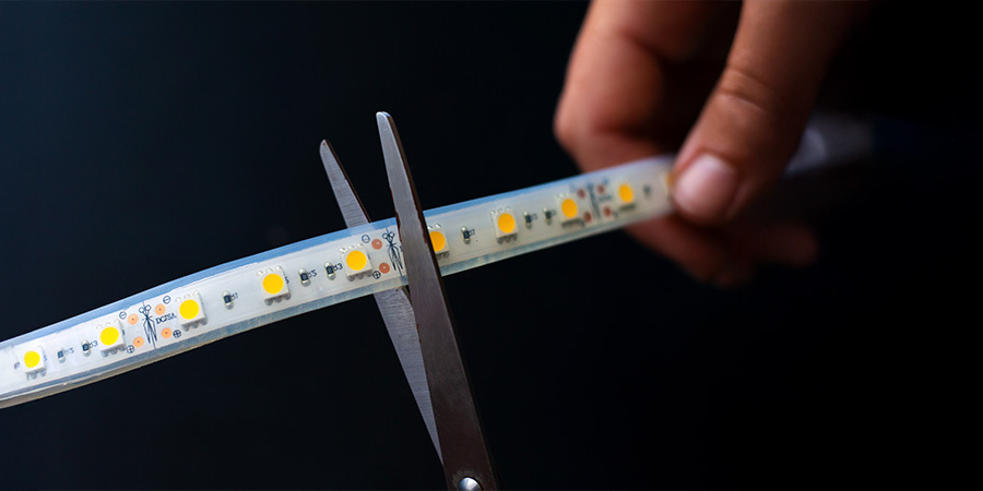 A close-up of a person using scissors to cut an LED strip at a marked cutting point, demonstrating easy customization for lighting projects.