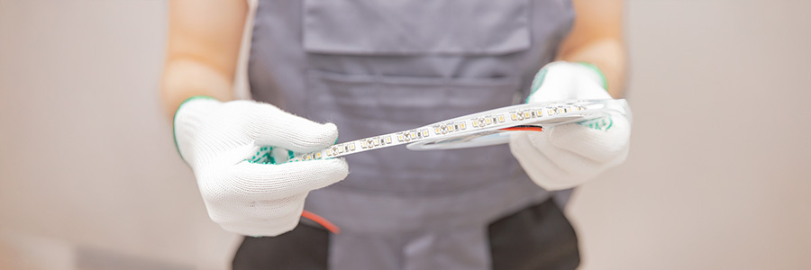 A close-up of a technician wearing gloves and holding a roll of LED strip light, showcasing the individual LED components and wiring.