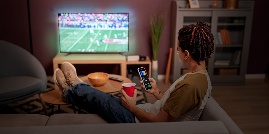 A young man relaxing on a couch, watching a football game on TV while holding a smartphone displaying the Leona Smart App